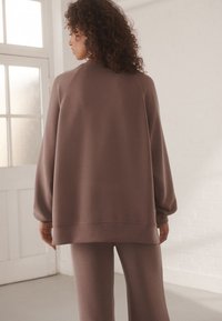 Felpa marrone oversize con maniche raglan, tessuto morbido e orlo a coste, abbinata a pantaloni jogger coordinati di una tonalità simile.