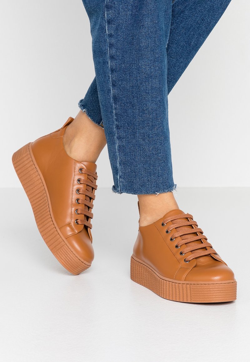 Baskets en simili cuir beige avec bout rond, semelle légère marron striée et lacets marron. Portées avec un jean bleu à longueur de cheville avec un ourlet effiloché.