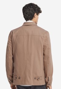 Brun bomullsjacka med en avslappnad passform, med en knappslå på ärmarna och en enkel kragsdesign. Slät struktur, utan mönster.