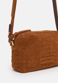 Braune Wildleder-Handtasche mit strukturiertem Gewebe, goldenem Reißverschluss und abnehmbarem Schultergurt. Mit einem Logodetail auf der Vorderseite.