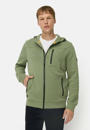 Jonge man die een lichtgroene hoodie met rits en zwarte ritssluitingzakken draagt, staand tegen een effen lichte achtergrond.