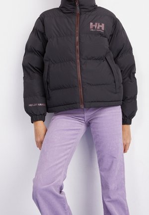 Person trägt eine schwarze Helly Hansen Daunenjacke mit Frontreißverschluss und hellvioletten Hosen vor einfachem Hintergrund.