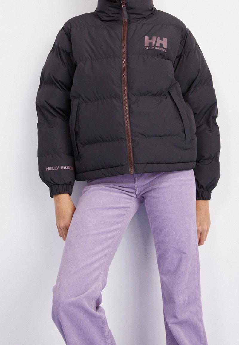 Personne portant une doudoune noire Helly Hansen avec une fermeture éclair à l’avant et un pantalon violet clair sur un fond uni.