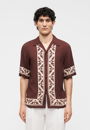 Άνδρας με σκούρα κατσαρά μαλλιά που φοράει πουκάμισο μανίκι κοντό σε μπορντό χρώμα με μπεζ ανθισμένο σχέδιο και λευκό παντελόνι, στέκεται μπροστά σε απλό φόντο.