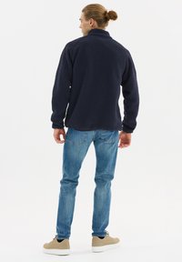 Laivastonsininen fleecetakki korkealla kauluksella, teksturoidulla kankaalla ja pitkillä hihoilla, yhdistettynä vaaleansinisiin farkkuihin ja vaaleisiin tennareihin.