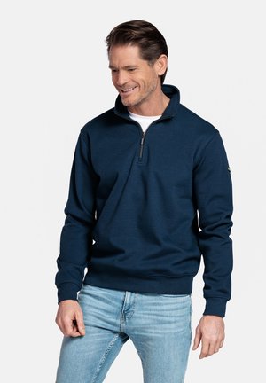 Man die een marineblauwe trui met een kwart-ritssluiting en lichtblauwe spijkerbroek draagt, glimlacht en naar links onder kijkt.