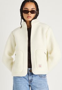 Giacca in pile crema con zip frontale, colletto alto, due tasche anteriori e dettagli in contrasto color tan, abbinata a un top scuro e jeans.