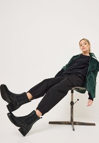 Botas de cuero negras hasta el tobillo con paneles laterales elásticos, tacón bloque y acabado texturizado. Combinadas con pantalones negros y una chaqueta verde.