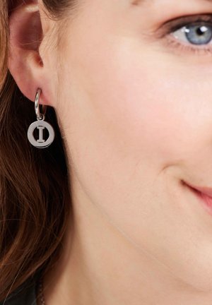 Silberner Creolenohrring mit einem runden Anhänger mit ausgeschnittenem Design, der knapp unter dem Ohrläppchen hängt und das Haar des Trägers ergänzt.