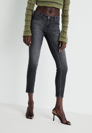 Strakzittende gewassen zwarte jeans gecombineerd met zwarte sandalen met hak en fijne bandjes, en een groene, opengewerkte cropped trui gedragen door een model met een donkere huid.