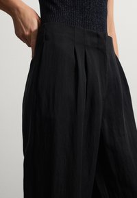 Pantaloni neri plissettati con una texture liscia, vita alta e tasche laterali, caratterizzati da pieghe sottili e una linea ampia.