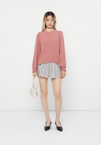 Rosa teksturert langermet topp med samlet skuldrer, paret med et grått mini-skjørt med folder. Modellen holder en liten hvit håndveske og har på seg svarte slingback-sko.