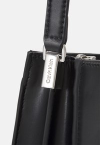 Sort læderhåndtaske med glat tekstur og struktureret design. Har sølvhardware og et mærke med "Calvin Klein."