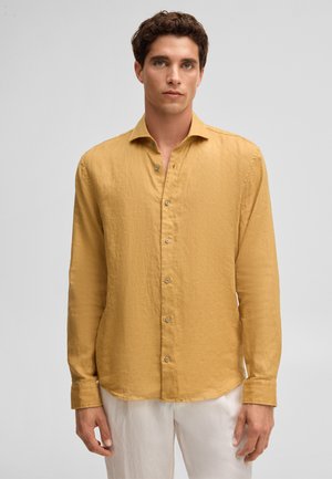 Young man with curly dark hair wearing a mustard yellow long-sleeve button-up shirt and white pants, facing forward.