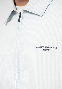 Ανοιχτό μπλε ριγέ φανέλα με φερμουάρ και γιακά, που διαθέτει το μαύρο κεντημένο λογότυπο "Armani Exchange Milano" στο στήθος.