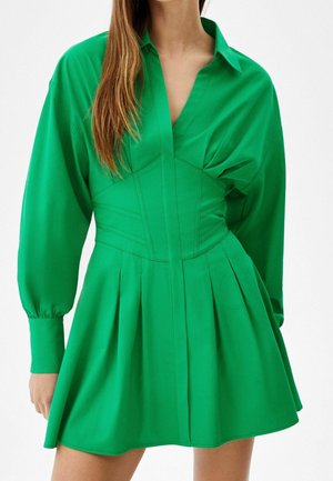 Donna che indossa un vestito verde brillante a maniche lunghe con vita aderente, gonna plissettata e collo aperto, in piedi davanti a uno sfondo semplice.
