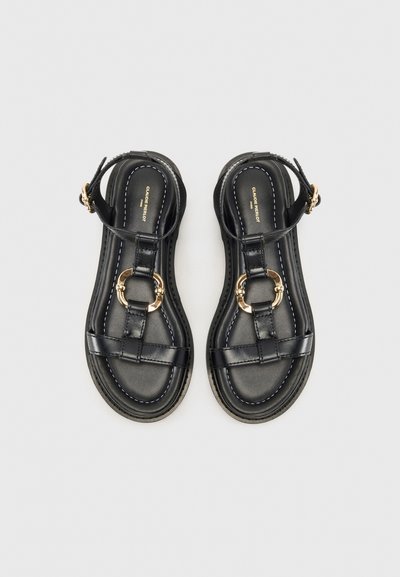 Sandales en cuir noir avec une semelle plate, dotées de lanières entrecroisées, d'une boucle décorative en ton or et de coutures blanches contrastantes.