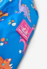 Vandeniui atsparus mėlynas lietpaltys su spalvingais dinozaurų ir gėlių raštais. Į komplektą įeina rožinė guminė etiketė su "JoJo splash splash" logotipu.