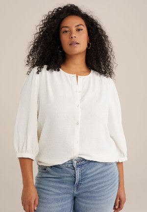 Witte, gestructureerde blouse met een knoopsluiting aan de voorkant en driekwart pofmouwen, gecombineerd met lichtblauwe spijkerbroeken.