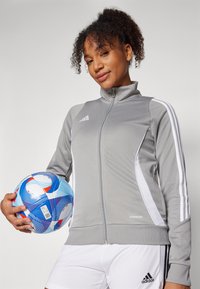 Gray zip-up athletic jacket with white stripes, made from smooth fabric. The model holds a blue and red patterned soccer ball.
