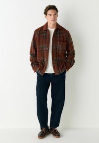 Brown plaid button-up shirt made of wool, with a chest pocket, paired with dark pants and brown shoes with a chunky sole.