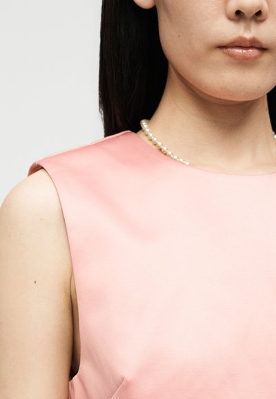 Γυναίκα που φοράει ένα ροζ φόρεμα χωρίς μανίκια και ένα κολιέ με πέρλες, απεικονίζεται από τον ώμο μέχρι λίγο κάτω από τα χείλη σε απλό φόντο.