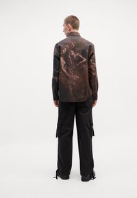 Chemise à manches longues en tissu sombre avec un imprimé texturé ; associée à un pantalon cargo noir avec des poches latérales et des zips. Des chaussures noires complètent la tenue.