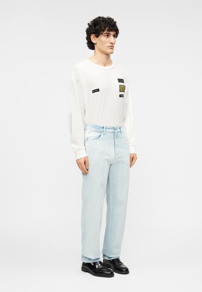 Λευκό πουκάμισο με μακριά μανίκια και τέσσερα μπαλώματα  στο μπροστινό μέρος, σε συνδυασμό με ανοιχτό μπλε τζιν με ίσια γραμμή και μαύρα παπούτσια, μινιμαλιστικός σχεδιασμός.