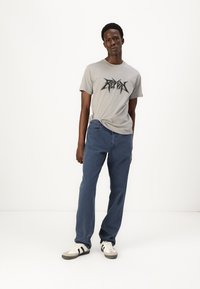 Grå t-shirt med sort grafisk design, parret med blå jeans. Sneakers med sorte striber, stående på en minimalistisch hvid baggrund.