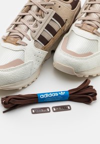 Tenisky Adidas v krémové a béžové barvě s texturovanými panely, s hnědými akcenty a tkaničkami. Součástí jsou také další hnědé tkaničky a plastové zámky na tkaničky.