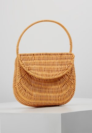 Sac à main en rotin tissé avec fermeture à rabat incurvée et une poignée arrondie, exposé sur un piédestal blanc sur un fond uni.