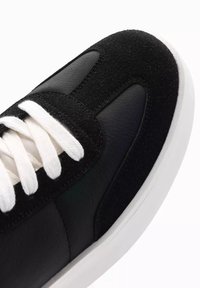 Svart sneaker med övre del i mocka och läder, vita snören, mjuk yta, rund form och en vit gummisul.