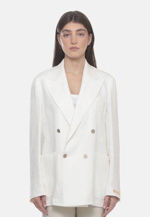 Donna con capelli lunghi e scuri che indossa un blazer bianco doppiopetto ampio con pantaloni beige su uno sfondo semplice.