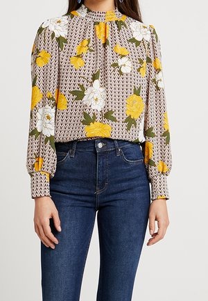 Donna indossa una camicetta a maniche lunghe con stampa floreale gialla e bianca e collo alto, abbinata a jeans a vita alta blu scuro.