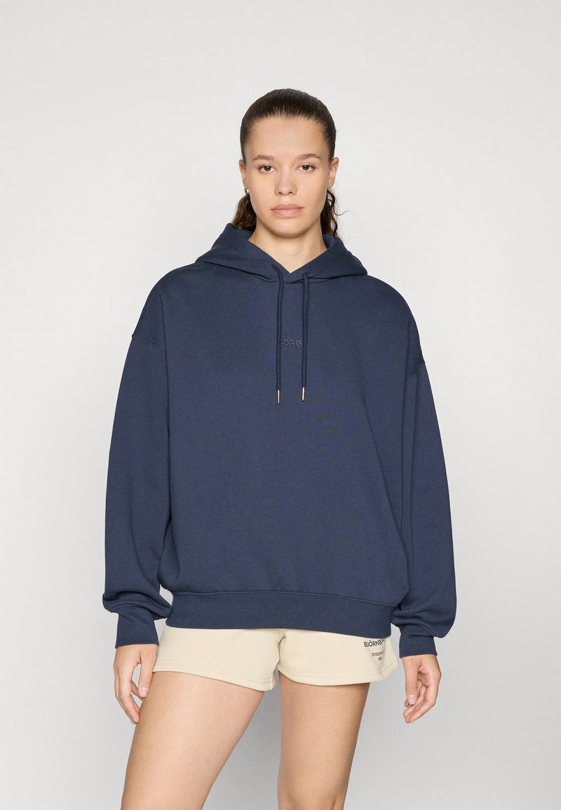 Felpa over-size blu navy in materiale morbido, con cappuccio spazioso, tasca frontale a marsupio e logo discreto sul petto. Abbinata a pantaloni corti beige.