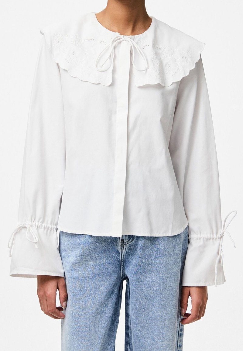 Blouse blanche avec un grand col brodé, noué au cou, et des manches resserrées avec des cordons. Tissu en coton lisse.