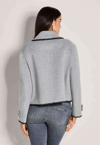 Vrouw met lang donker haar, gekleed in een grijze cropped jasje met zwarte accenten en grijze slim-fit jeans, staand met haar rug naar de camera tegen een effen achtergrond.