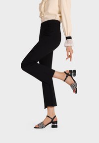 Sandalini neri con un tacco spesso presentano intricate decorazioni in argento e un design con cinturini, abbinati a pantaloni neri attillati e una blusa crema.