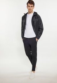 Sort letvægtsjakke med hætte, der har to frontlommer, en hvid t-shirt under, sorte bukser og hvide sneakers.