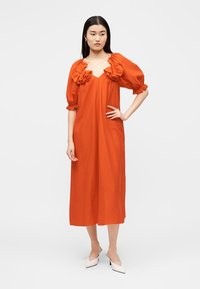 Γυναίκα στέκεται με το δεξί χέρι στη μέση, φορώντας ένα φαρδύ πορτοκαλί μεσαίο φόρεμα με φουσκωτά μανίκια και λεπτομέρειες με βολάν στο ντεκολτέ.
