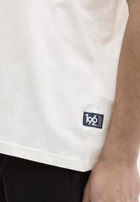 Valkoinen puuvillainen t-paita, jossa on pieni tummansininen etiketti, jossa lukee numero "196" alahelmassa. Yksinkertainen muotoilu ja rento istuvuus.
