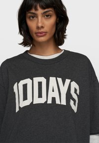 Dunkelgraues, oversized T-Shirt mit fettem weißen Text "100 TAGE." Der Stoff wirkt weich und hat einen runden Halsausschnitt. Eine weißes Unterhemd ist sichtbar.
