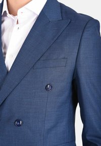 Blazer blu doppiopetto in tessuto texturizzato, con rever a lancia e due bottoni scuri. Dettaglio della pochette sul petto.