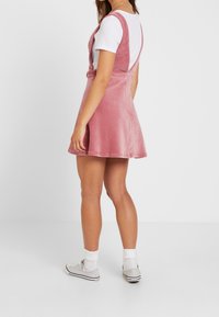 Roze corduroy korte jurk met verstelbare bandjes, A-lijn rok en een lage rugontwerp, gecombineerd met een wit T-shirt en grijze sneakers.