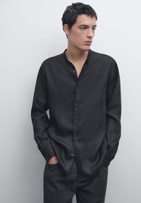 Zwarte langemouwen shirt met een bandkraag en knoopsluiting aan de voorkant, gemaakt van textuurstof. Draagt het met donkere jeans, handen in de zakken.
