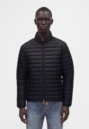 Mand iført sort vatteret jakke med orange lynlås og blå jeans, stående mod en ensfarvet hvid baggrund.