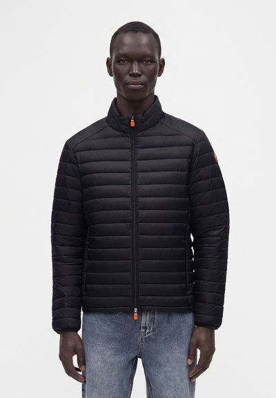 Un homme porte une veste matelassée noire avec une tirette de fermeture éclair orange et un jean bleu, debout devant un fond blanc uni.