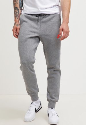 Mężczyzna w szarych spodniach dresowych z czerwonym logo na udzie, białych skarpetkach Nike i białych butach Nike stoi na jednolitym tle.