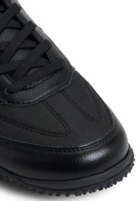 Zapatilla negra con parte superior de cuero texturizado y nylon, punta redondeada, cordones planos y suela de goma moldeada con pequeños pinchos para tracción.