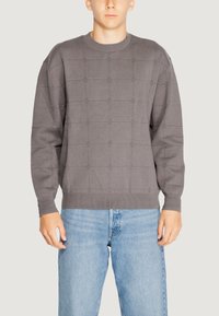 Maglione grigio in maglia con un motivo a griglia testurizzato, polsini e scollatura a coste, abbinato a jeans azzurri chiari.